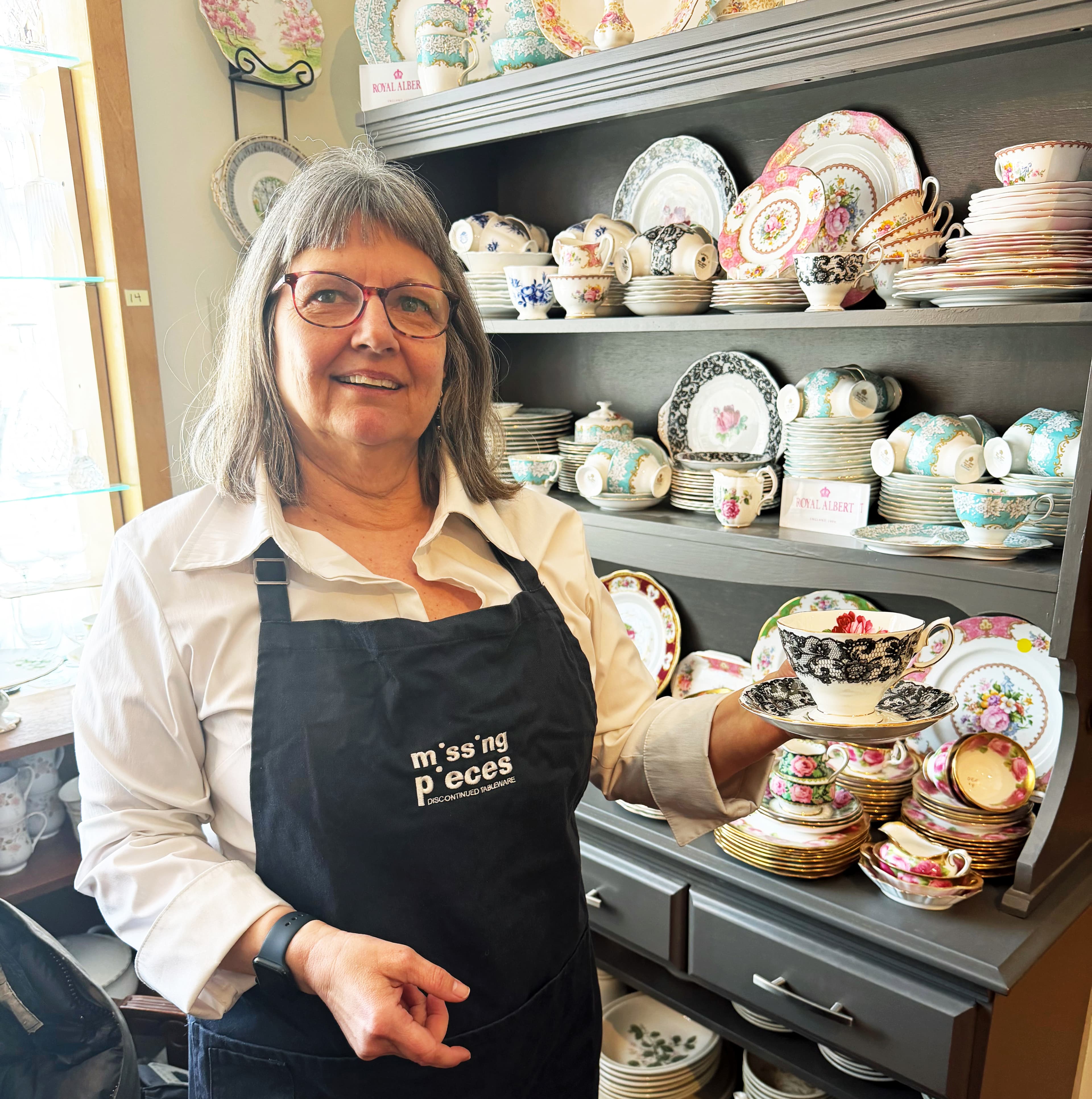 Janice Perkins in her Missing Pieces shop, holding a teacup in front of shelves of fine china